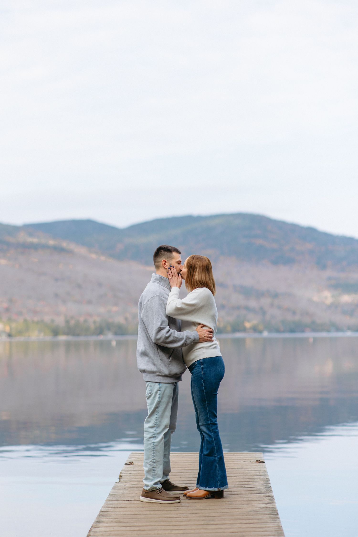 A couple stands together on a wooden dock overlooking a serene lake with mountains in the background.