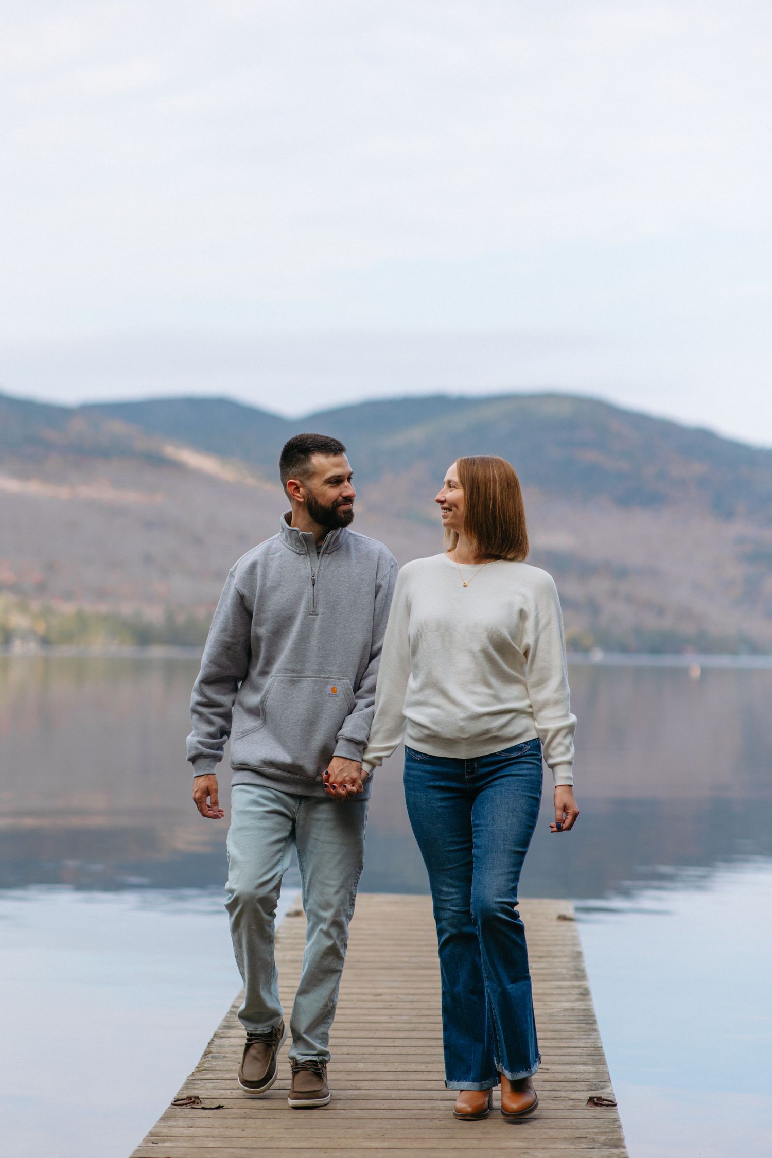 A couple strolls hand in hand on a lakeside dock with mountains and autumn colors in the background.