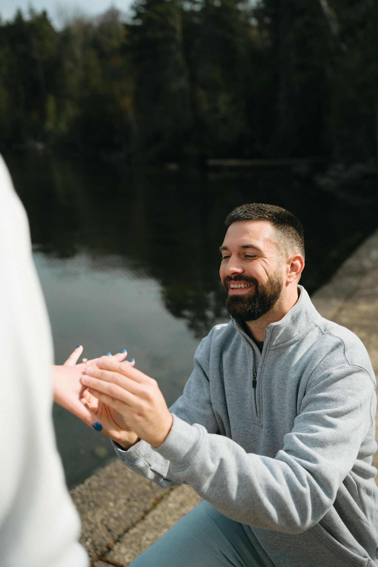 A couple shares a joyful moment by a lake, with one person laughing while wearing a grey sweatshirt.