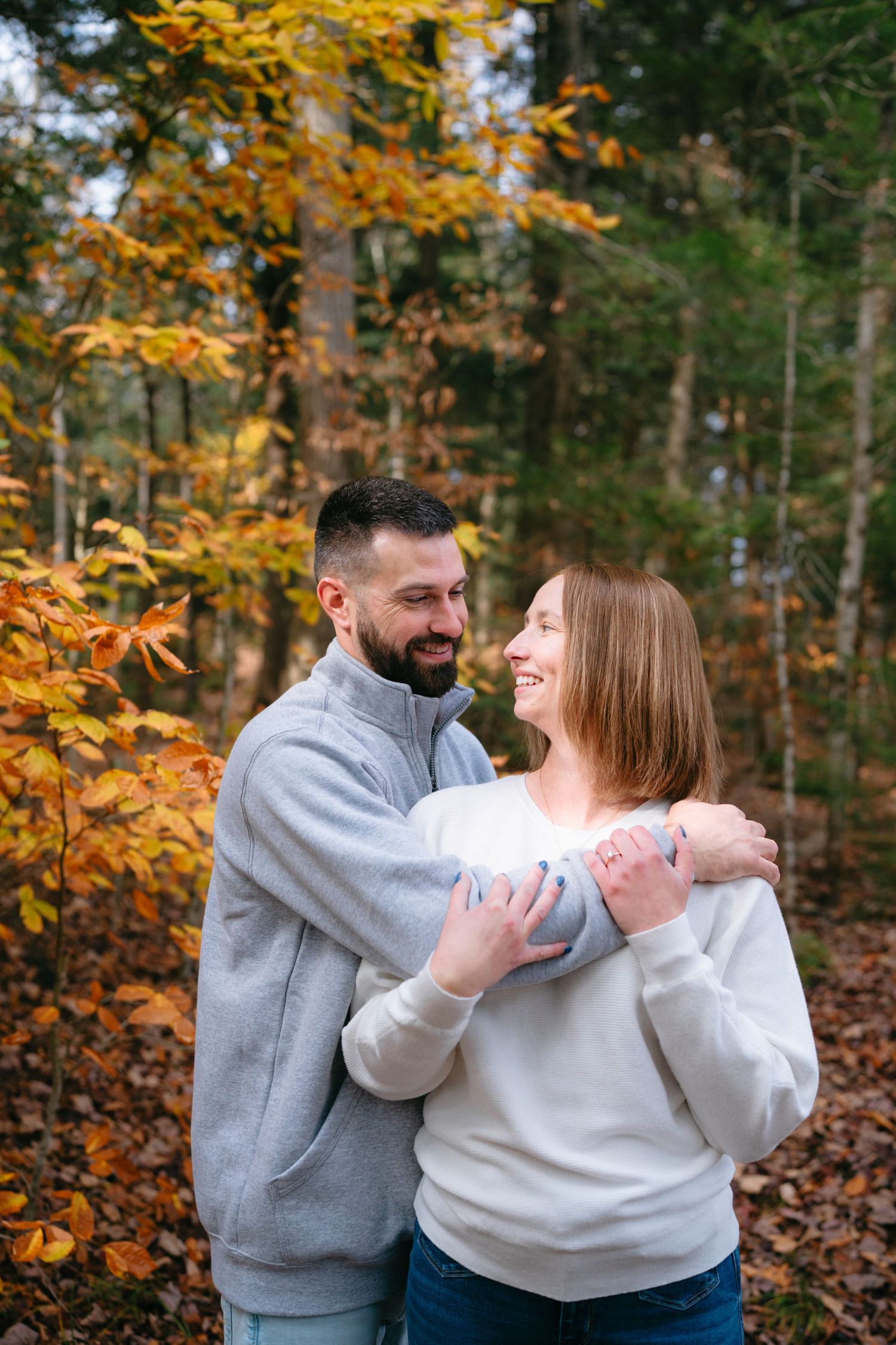 A couple wearing matching gray and white sweaters share tender moments in an autumn forest with golden leaves.