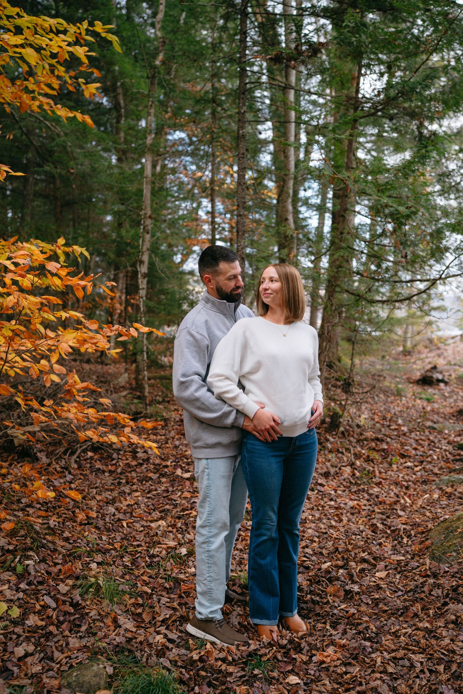 A couple embraces while standing among fallen autumn leaves in a forest with golden foliage.