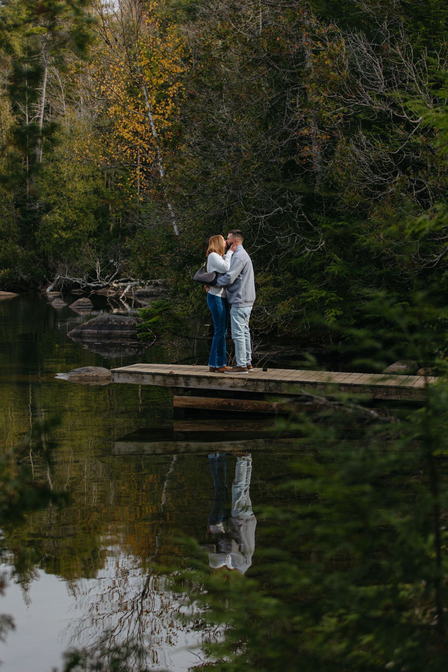 A couple embraces on a wooden dock by a serene lake surrounded by autumn trees reflecting in the calm water.