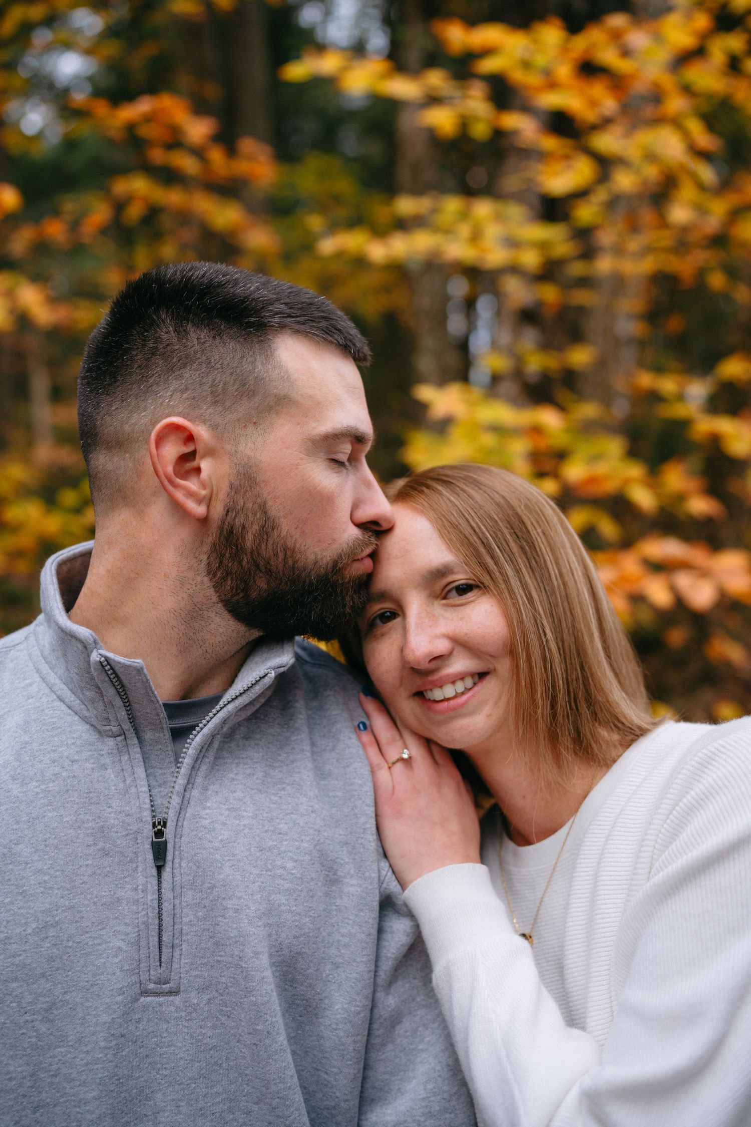 A couple in matching sweaters share an intimate moment against vibrant autumn foliage in the woods.