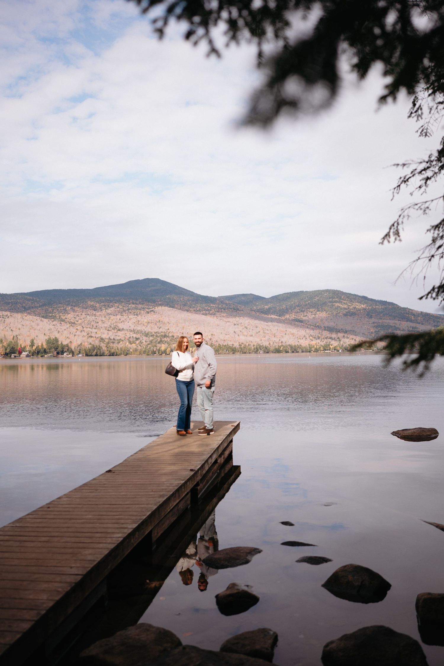 A wooden dock extends over a serene lake with mountains in the background on a cloudy autumn day.