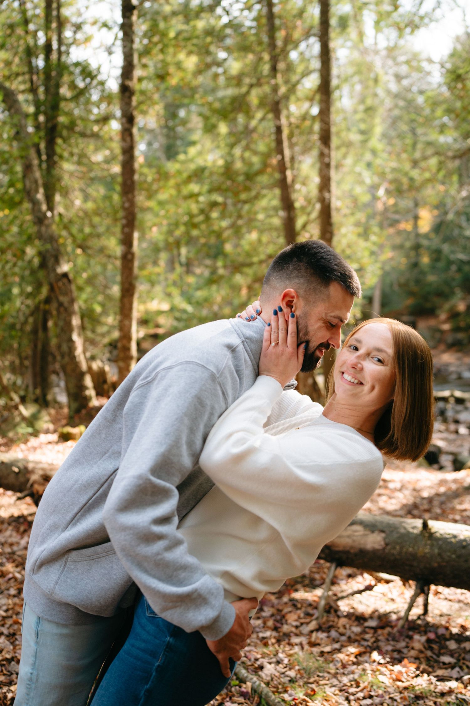 A playful couple in casual wear embrace and laugh together in a sunlit forest setting.