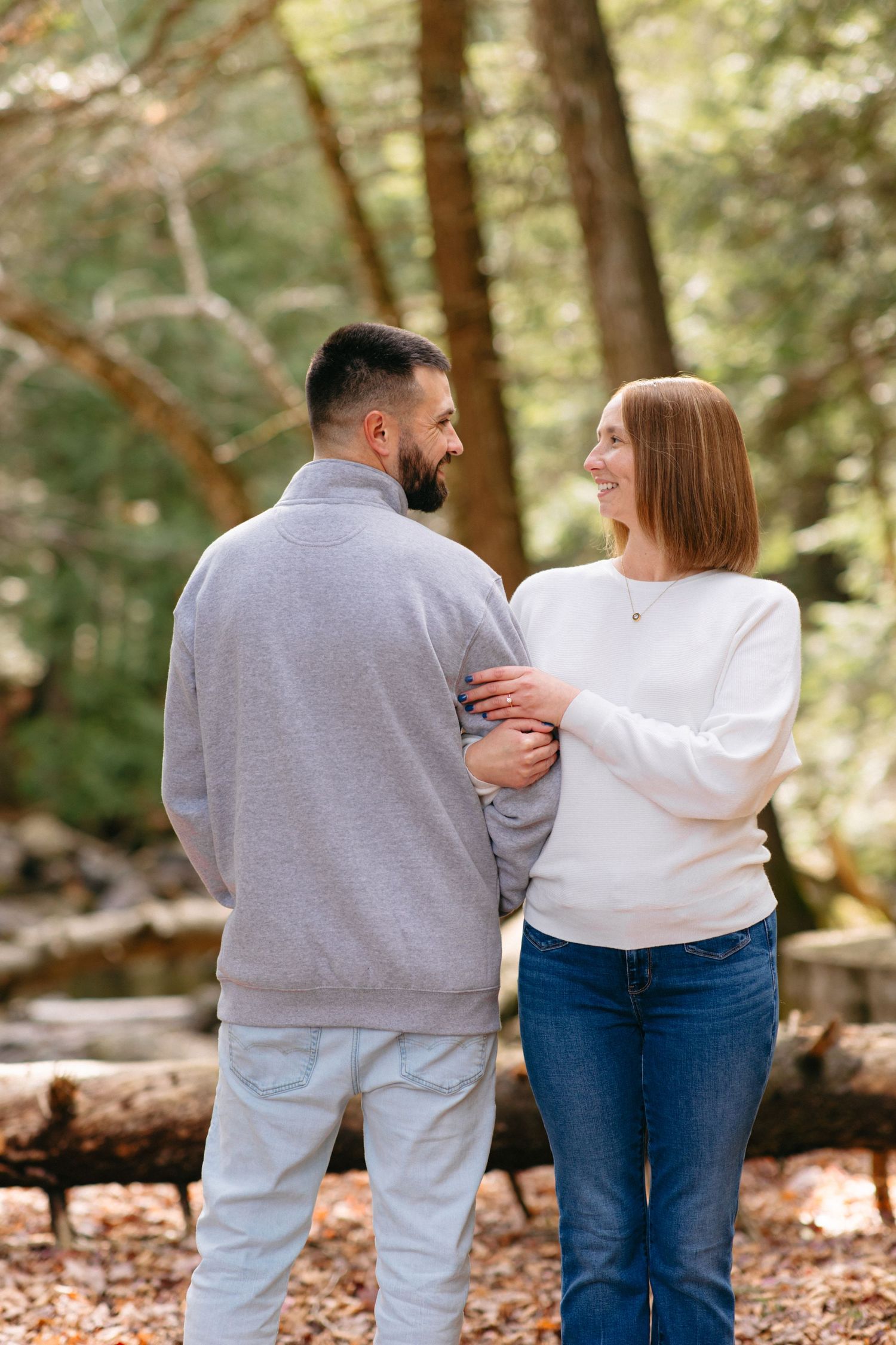A couple in casual attire walks together through a wooded area with fallen autumn leaves.