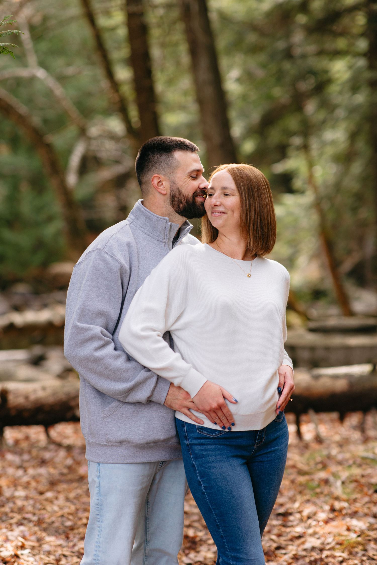 A couple in coordinating gray and white sweaters share a tender moment among fallen leaves in the woods.