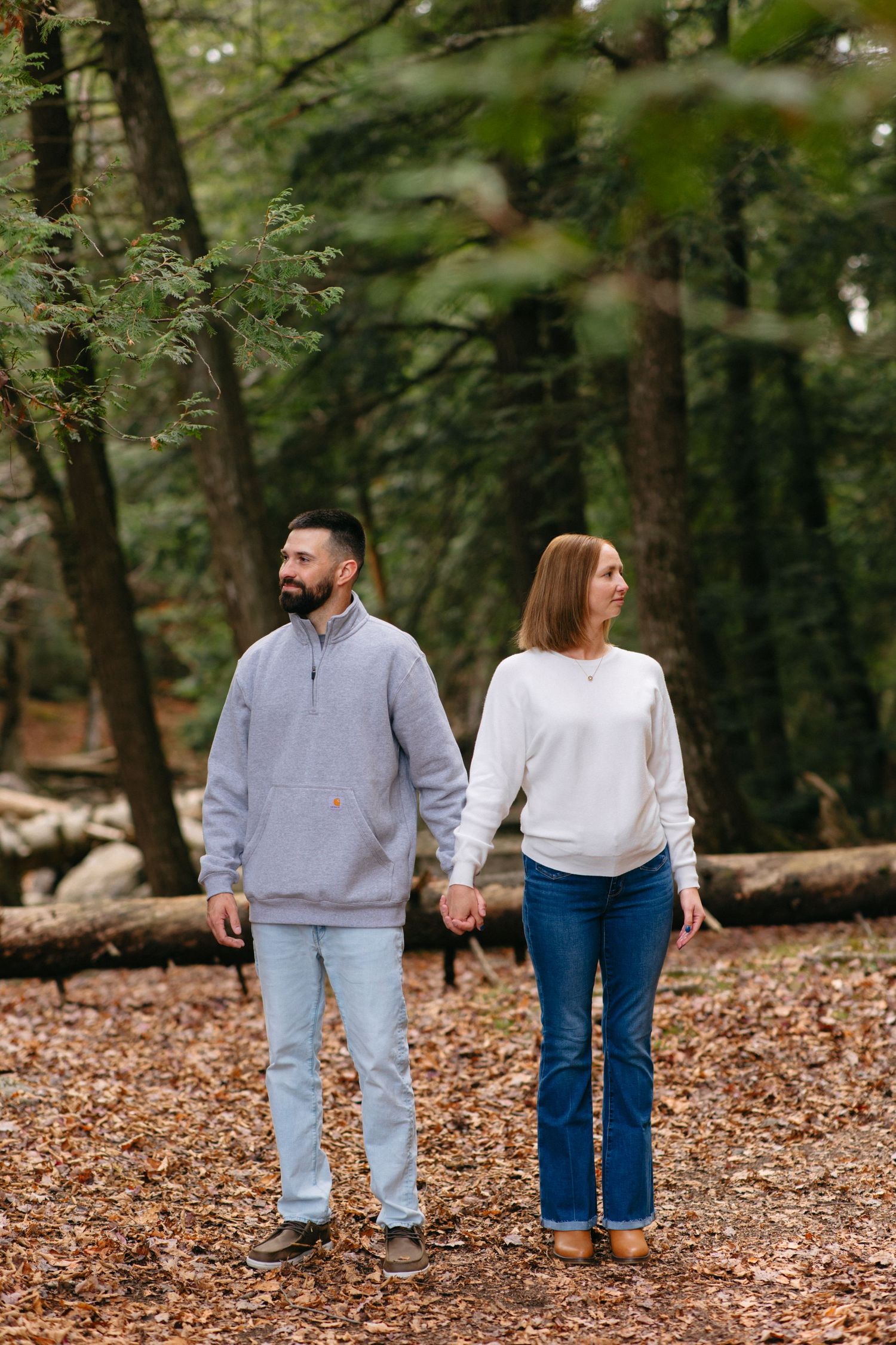 A couple takes a stroll through fallen leaves in a wooded area wearing matching casual outfits.