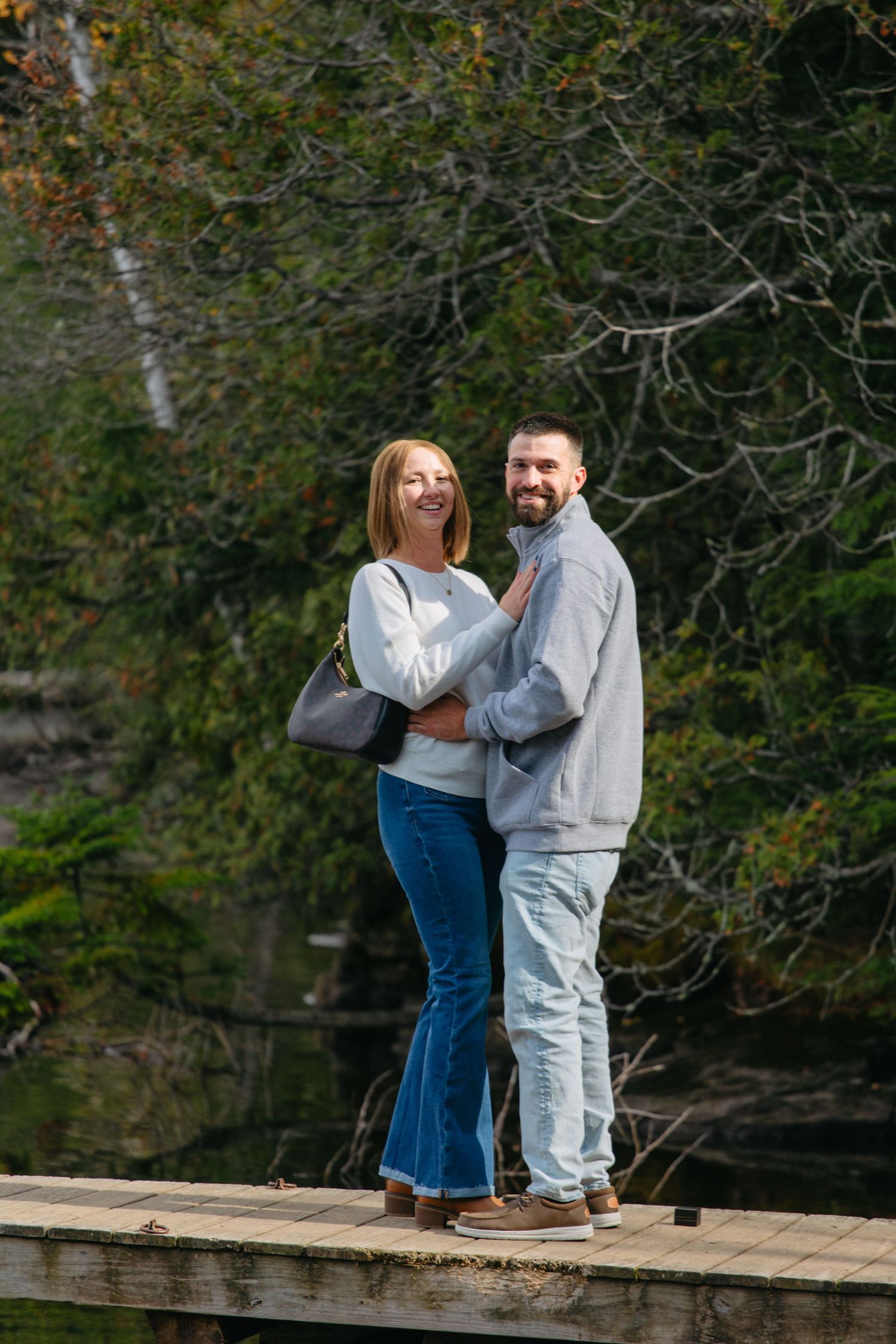 A couple stands together on a rustic wooden dock surrounded by fall foliage and calm waters.