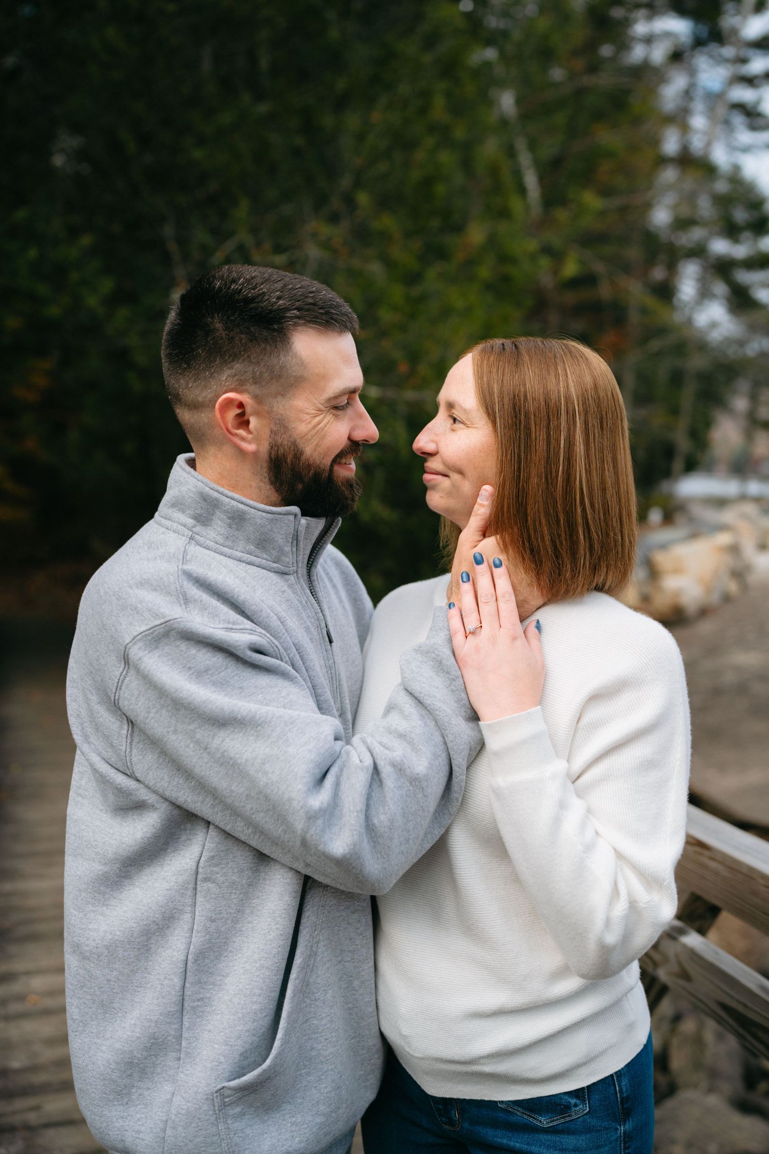A couple in matching sweaters share a loving moment on a wooden bridge surrounded by evergreen trees.