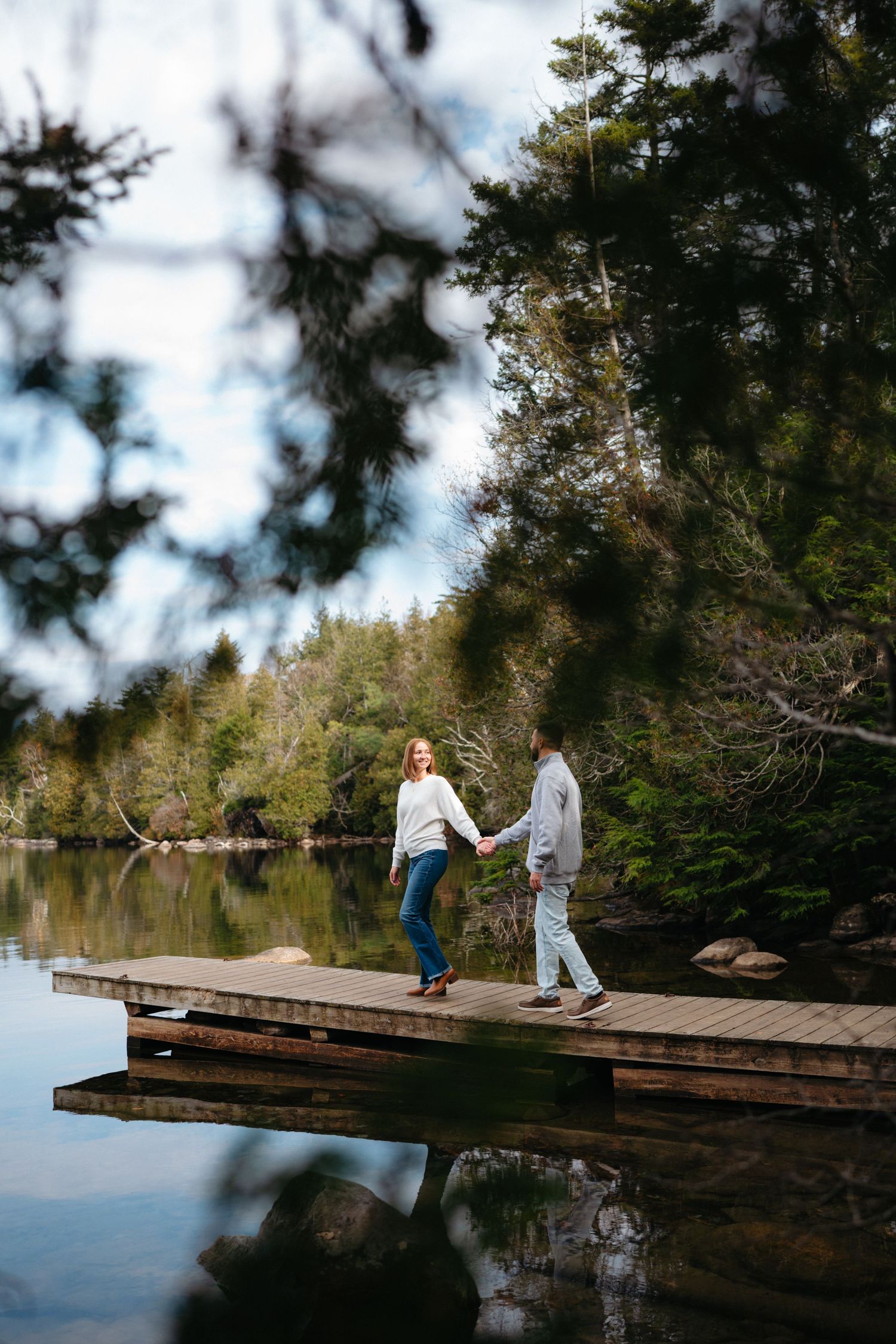 A couple walks along a wooden dock surrounded by evergreen trees and reflective water on a cloudy day.