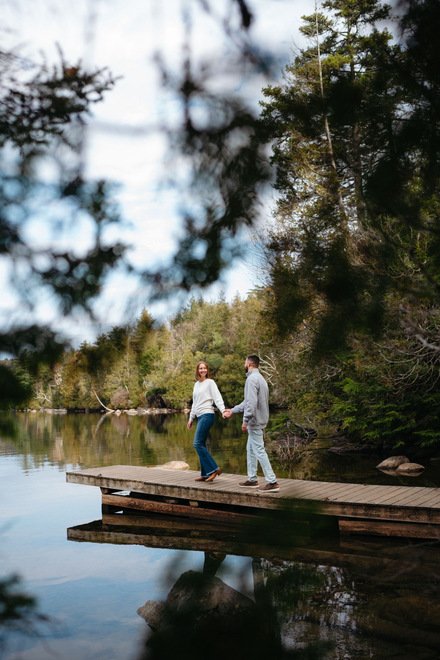 Two people walk along a wooden dock overlooking a peaceful lake surrounded by evergreen trees.