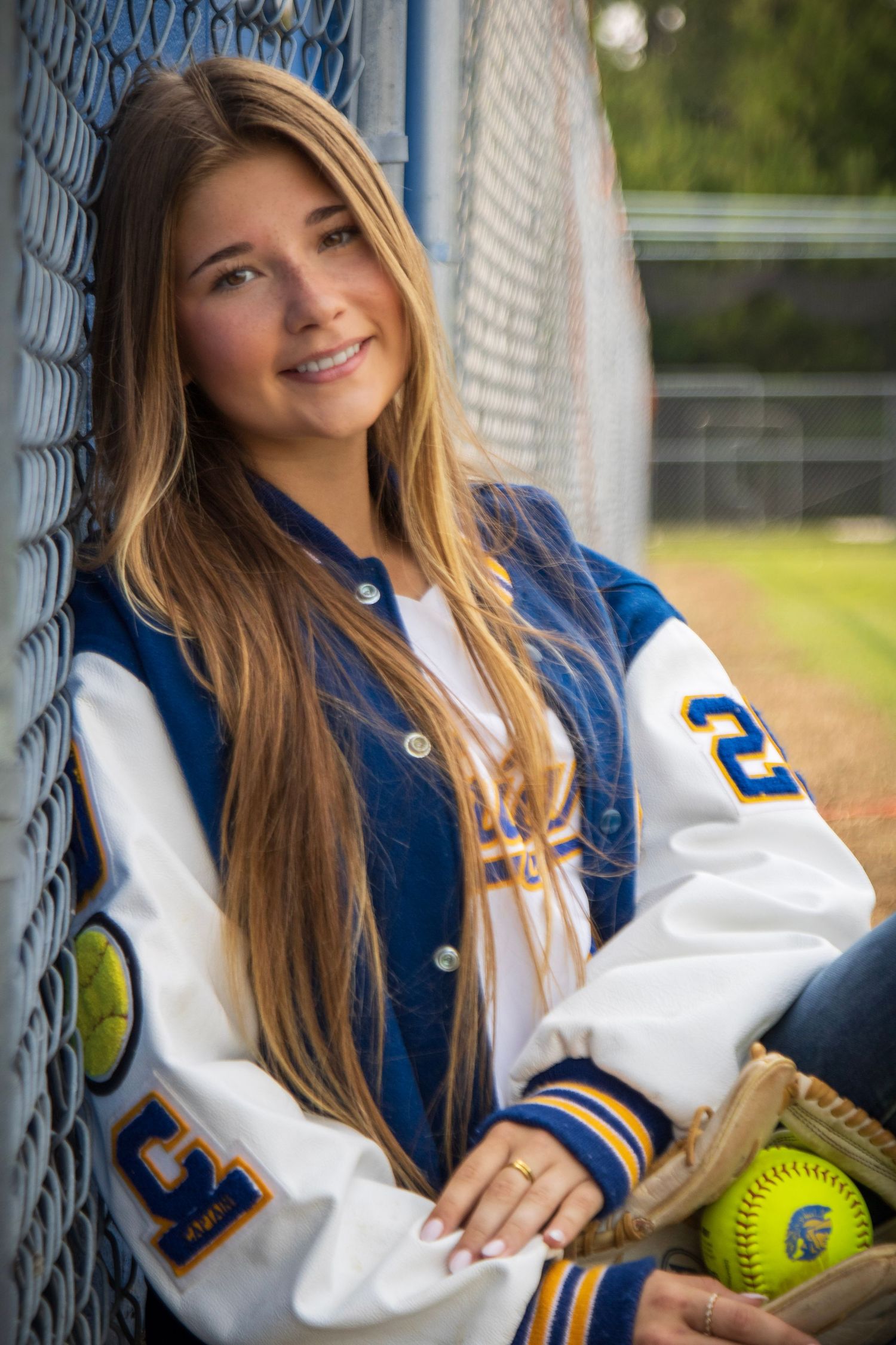 High school softball player in navy and white varsity jacket poses with yellow softball against chain link fence.