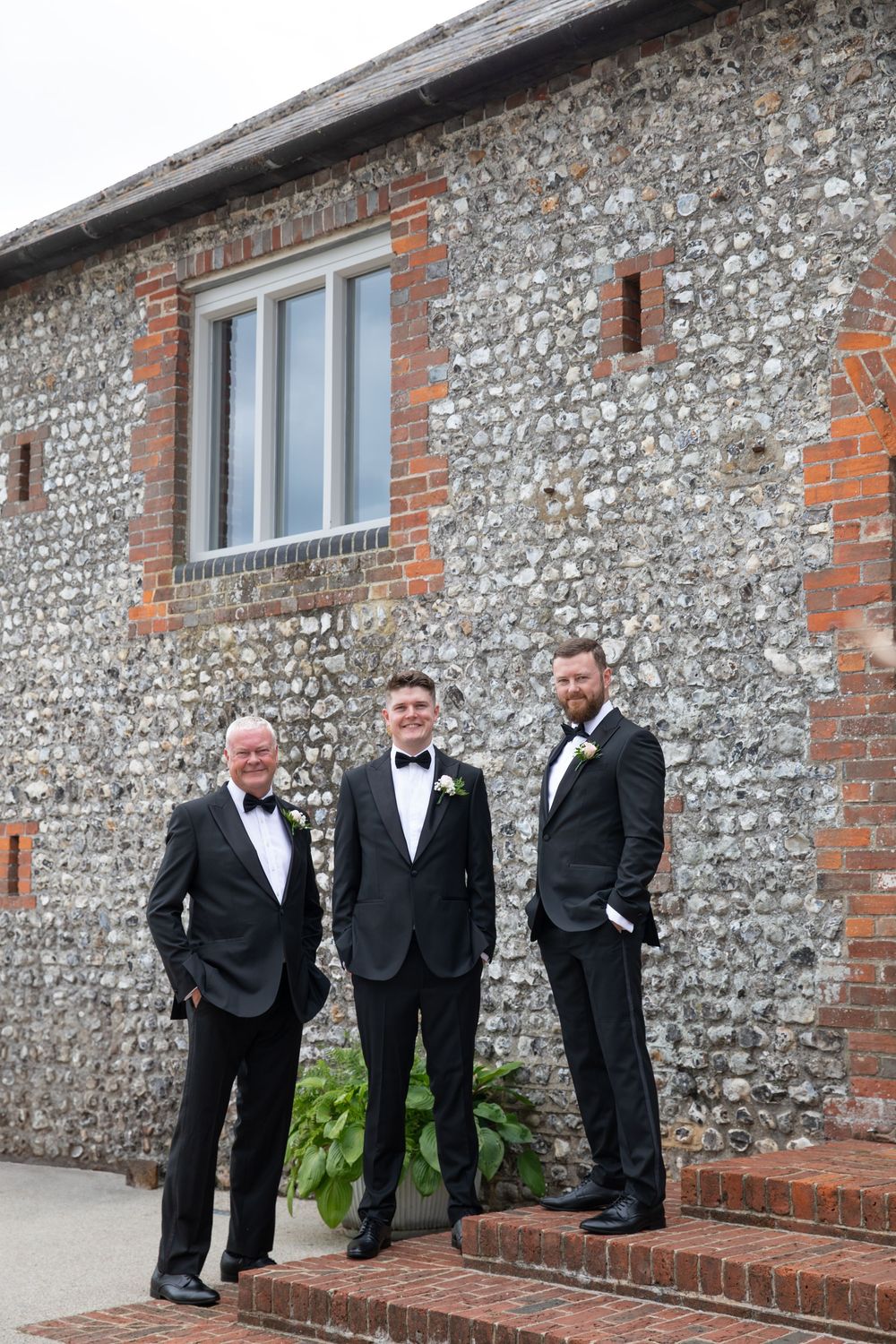 Groomsmen in black tuxedos pose casually against a rustic stone wall.