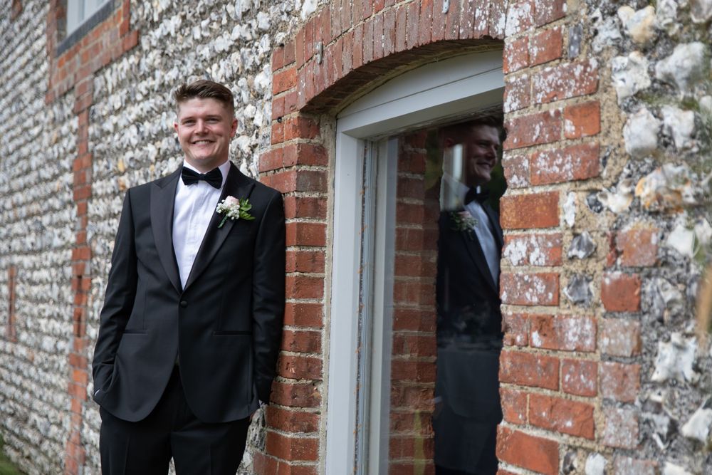Classic groom in black tuxedo poses against rustic brick and stone wall.