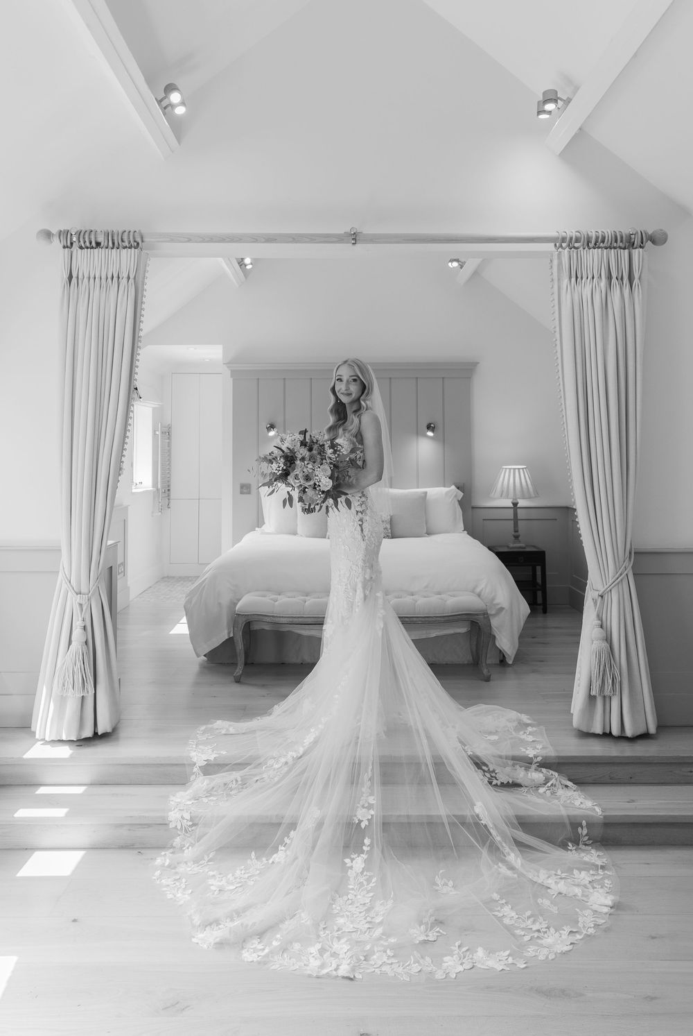 Elegant white wedding dress with long lace train displayed in bright, airy room with white curtains.