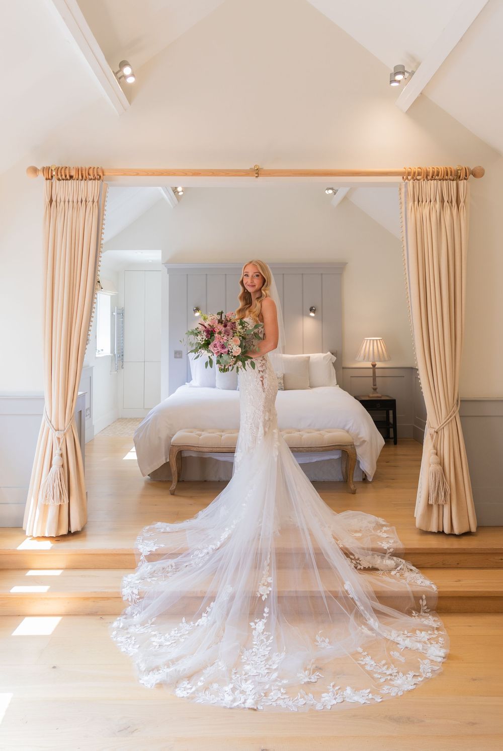 An elegant bridal suite with a flowing lace wedding dress and train displayed against cream curtains and wooden floors.
