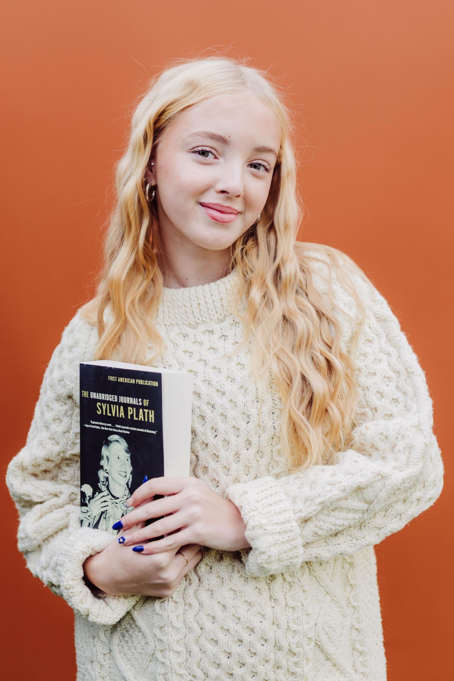 Person wearing cream cable knit sweater holds book against orange background in creative photo series.