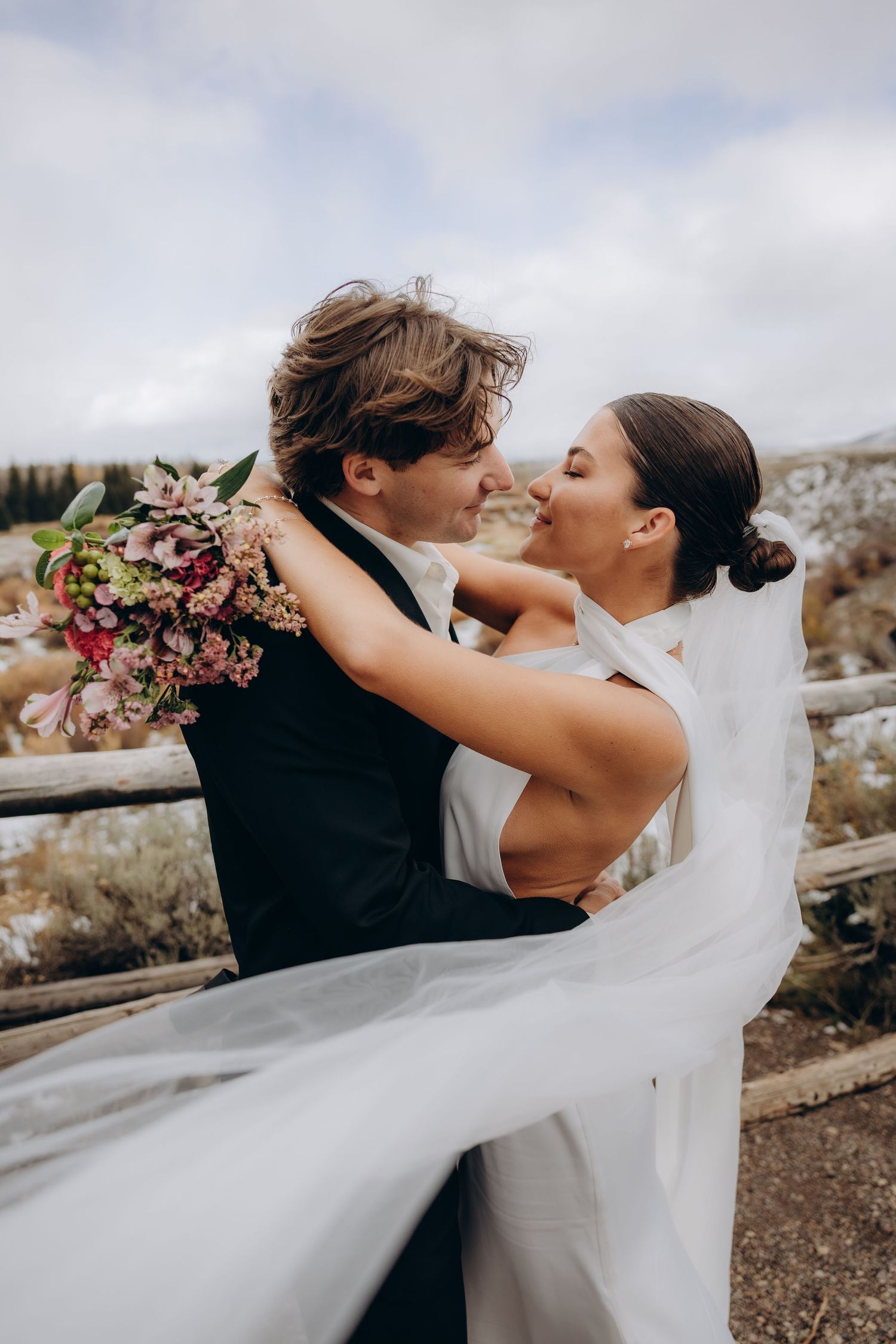 A romantic wedding couple embraces and dances together outdoors with a flowing white veil catching the wind.