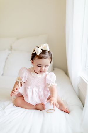 Baby wearing pink smocked dress plays on white bedding near window.