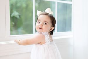 Baby in white dress sits by bright window with natural lighting.