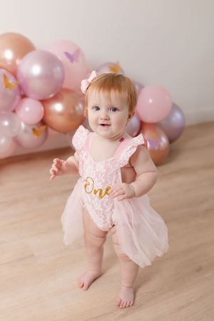 Baby in pink lace romper crawling on wooden floor with pink balloons in background.