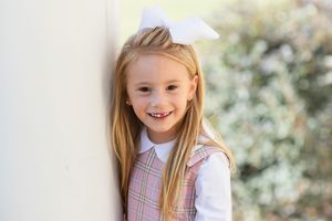 Child in white dress smiling outdoors with blurred background.