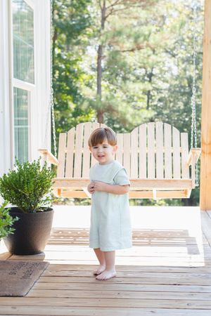 Child in white outfit exploring wooden porch with potted plants.