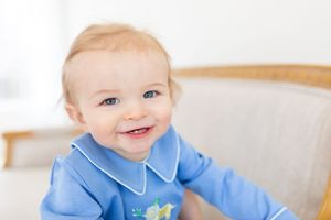 Baby wearing blue collared outfit smiling on white bedding.