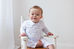 Baby wearing white patterned outfit sitting in wooden chair.
