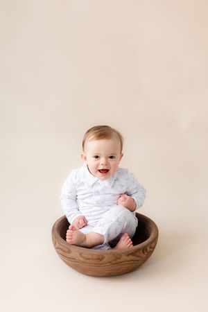 Baby in white outfit sitting in wooden bowl against neutral backdrop.