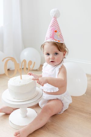 Baby in white outfit and pink party hat sitting with white birthday cake.