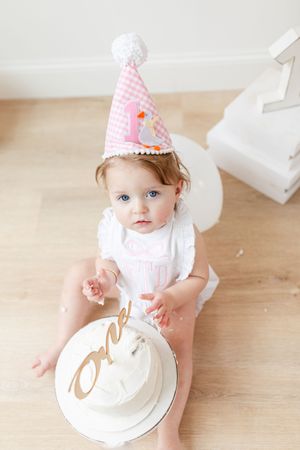 Baby wearing party hat reaching for white birthday cake on wooden floor.