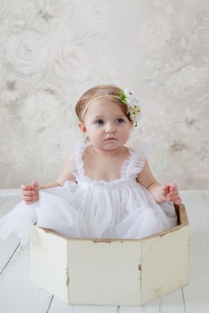 Sweet baby in white dress poses in vintage wooden box against textured backdrop.