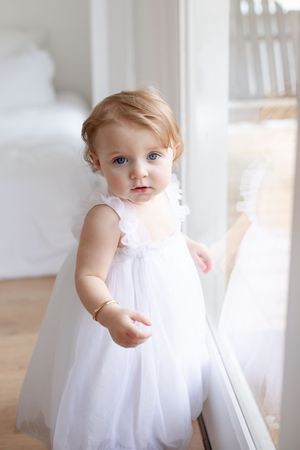 Baby wearing flowing white dress twirls in bright indoor setting.