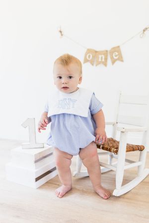 Baby in blue gingham outfit next to white wooden rocking horse.