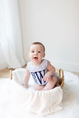 Baby in white and navy outfit sits in golden basket against white background.