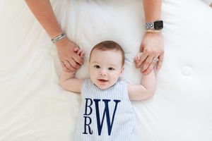 Baby wearing monogrammed outfit lies on white bedding with hands visible.