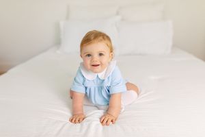 Baby wearing light blue outfit lying on white bedding in bright natural light.