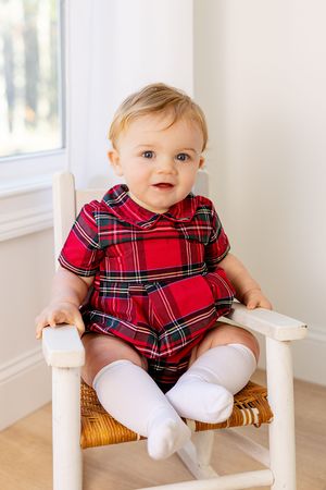 Baby in red plaid holiday dress and white tights sitting on woven chair.