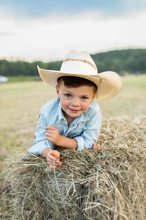 A child in a cowboy hat lies playfully on a hay bale in a sunny field.
