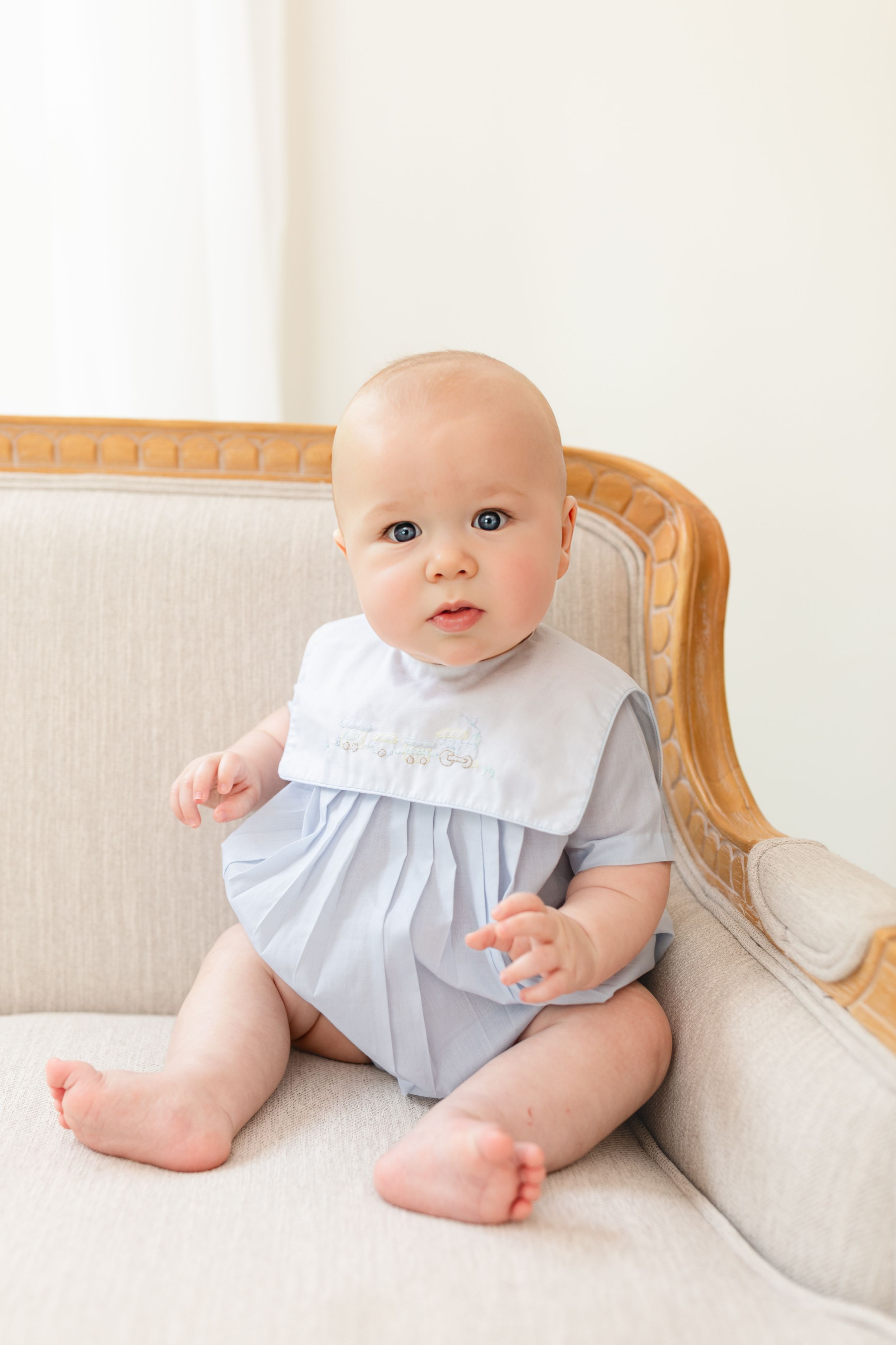 Adorable baby in light blue romper with bib sitting on cream French chair, looking curiously at camera.