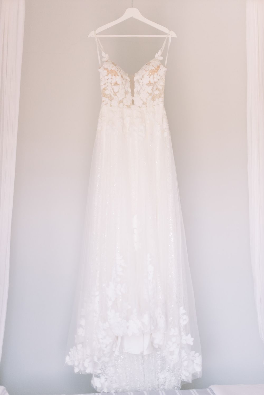 White lace wedding dress with floral appliques hangs elegantly on a white hanger against a light gray wall.