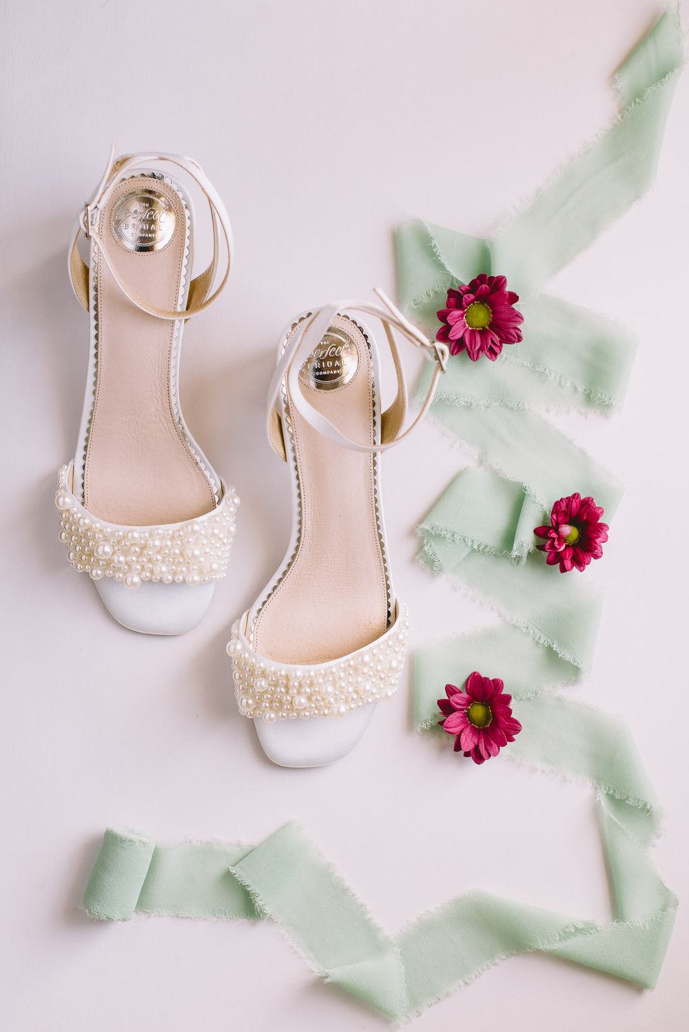White pearl-embellished bridal shoes with mint green ribbons and pink flowers arranged on a light background.