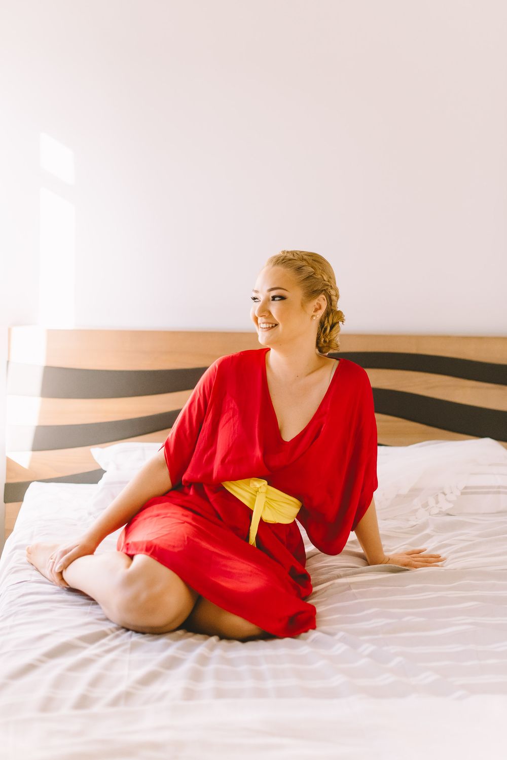 Woman in red dress lounges gracefully on white bedding in a bright, minimalist bedroom setting.