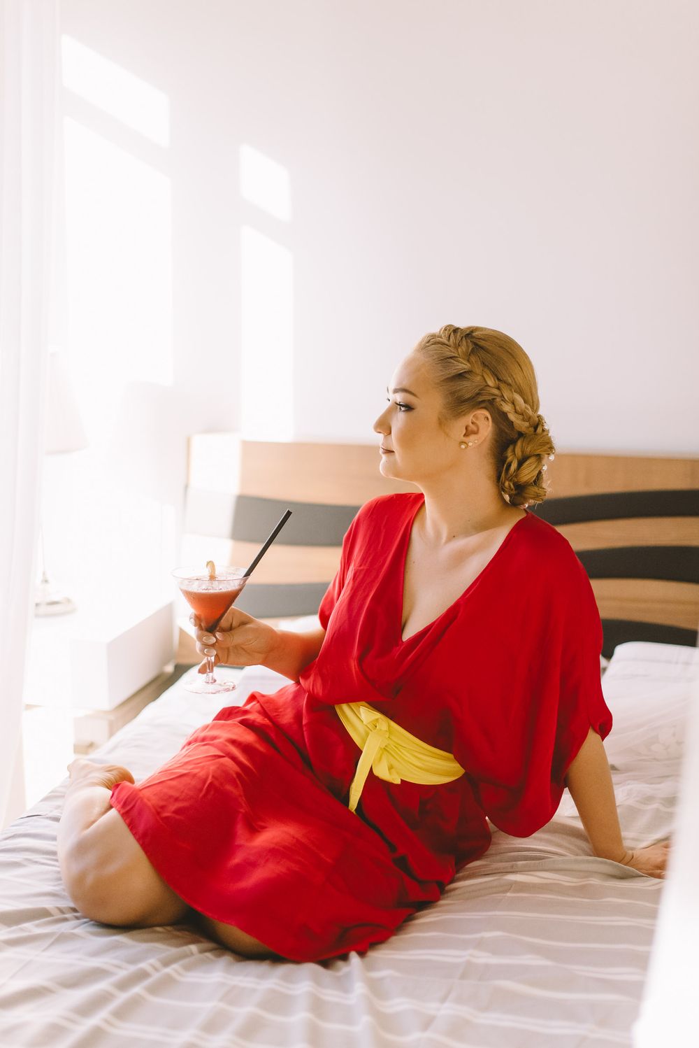 Person in a vibrant red dress lounges gracefully on white bedding while holding a drink.