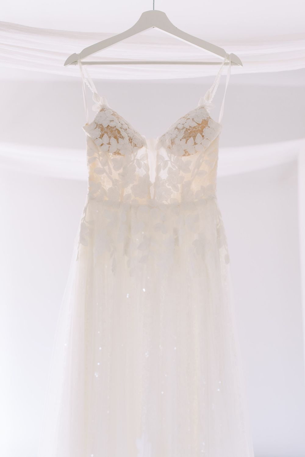 A delicate white wedding dress with floral lace appliques hangs elegantly against a light background.