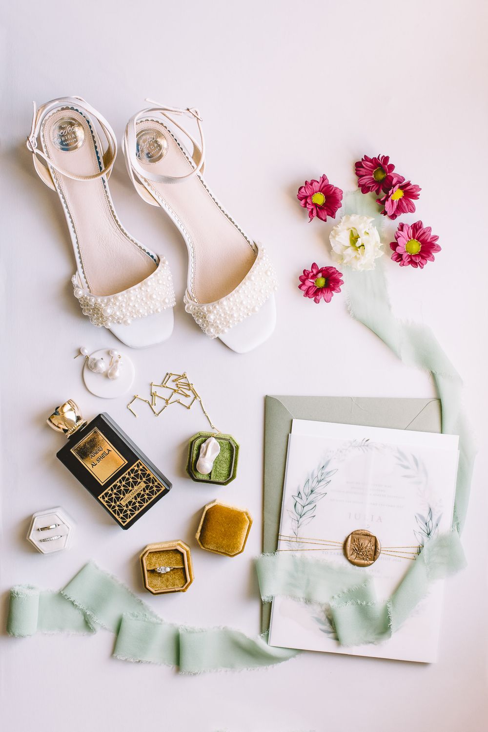 Elegant white bridal shoes, pink flowers, and wedding stationery with sage green accents arranged on white surface.