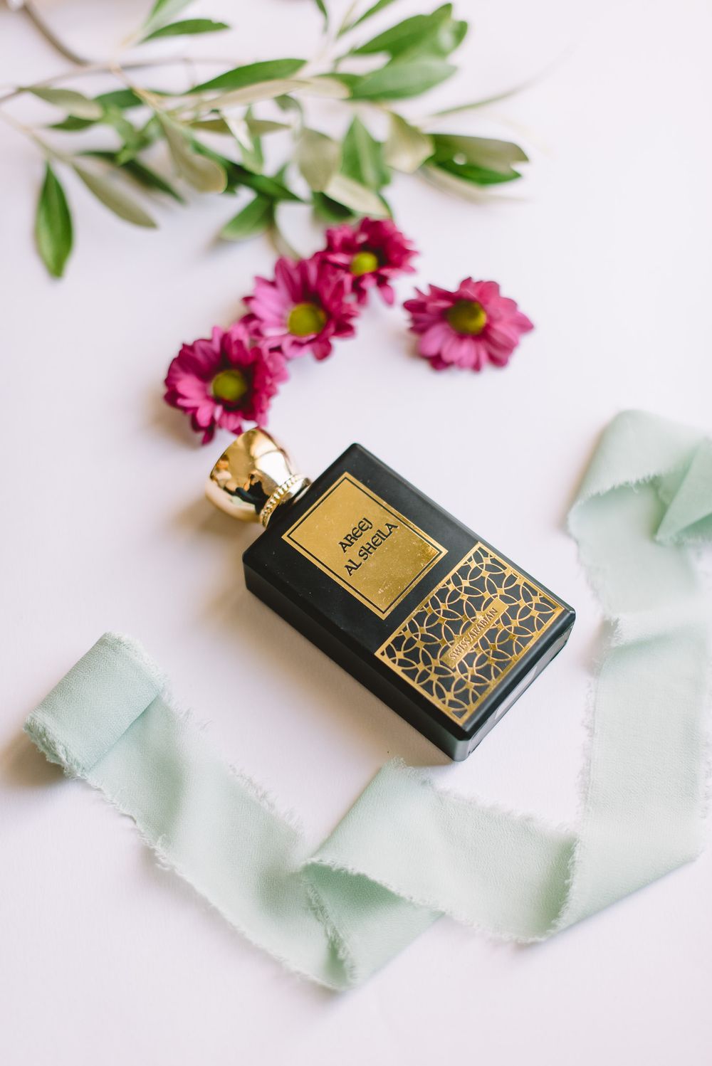 Black and gold perfume bottle on white surface with pink flowers and green leaves arranged artistically.