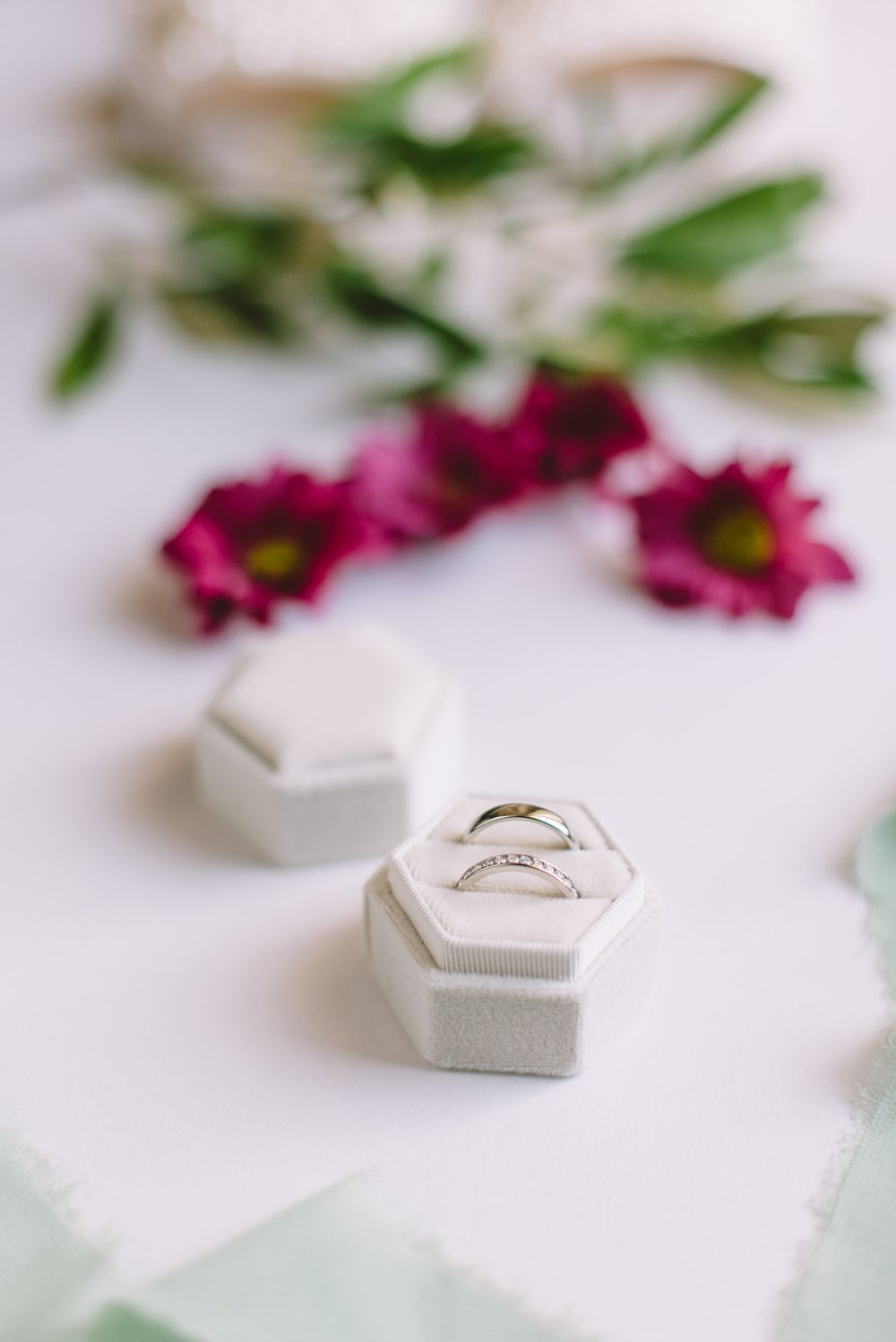 Close-up of delicate wedding rings in white hexagonal ring box with pink flowers in soft focus.